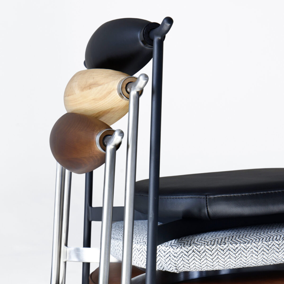 06_ASSC-CR-ASBL-01 A side view of stacked modern chairs with differently coloured wooden backrests—black, natural wood, and dark brown—and various textured seat cushions, against a plain white background.