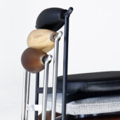 A side view of stacked modern chairs with differently coloured wooden backrests—black, natural wood, and dark brown—and various textured seat cushions, against a plain white background.