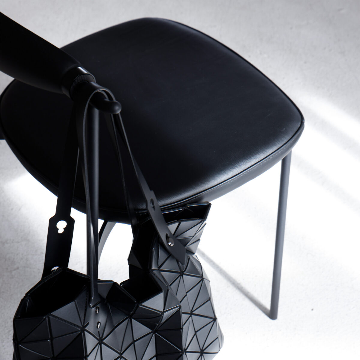 07_ASSC-CR-ASBL-01 A black geometric-patterned handbag hangs on the backrest of a black, minimalist chair with thin metal legs, set on a light-coloured floor with sunlight casting shadows.