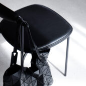 A black geometric-patterned handbag hangs on the backrest of a black, minimalist chair with thin metal legs, set on a light-coloured floor with sunlight casting shadows.