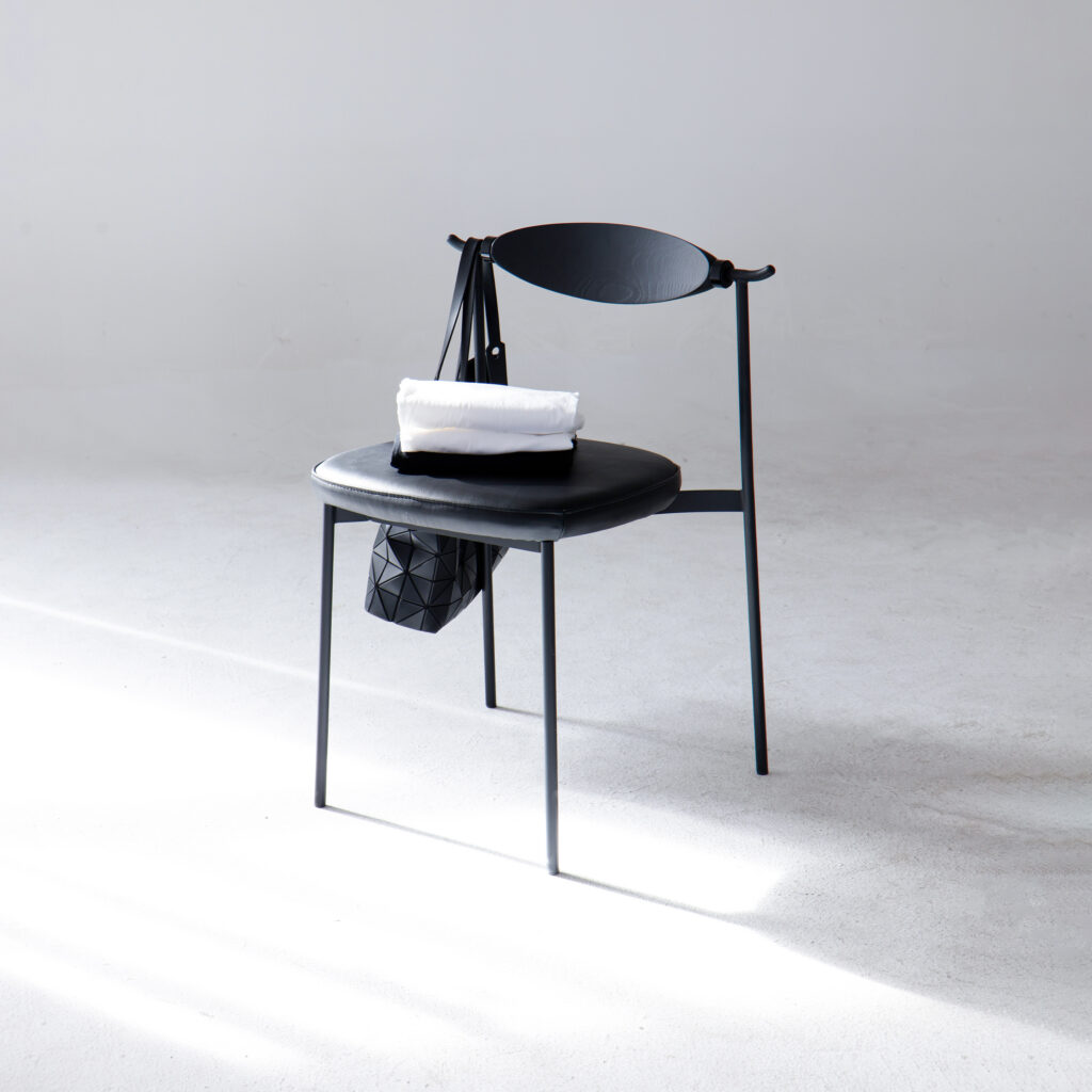 A modern black chair with a curved backrest holds a folded white towel and a black geometric-patterned bag, set in a minimal, bright space with a light grey floor and wall.