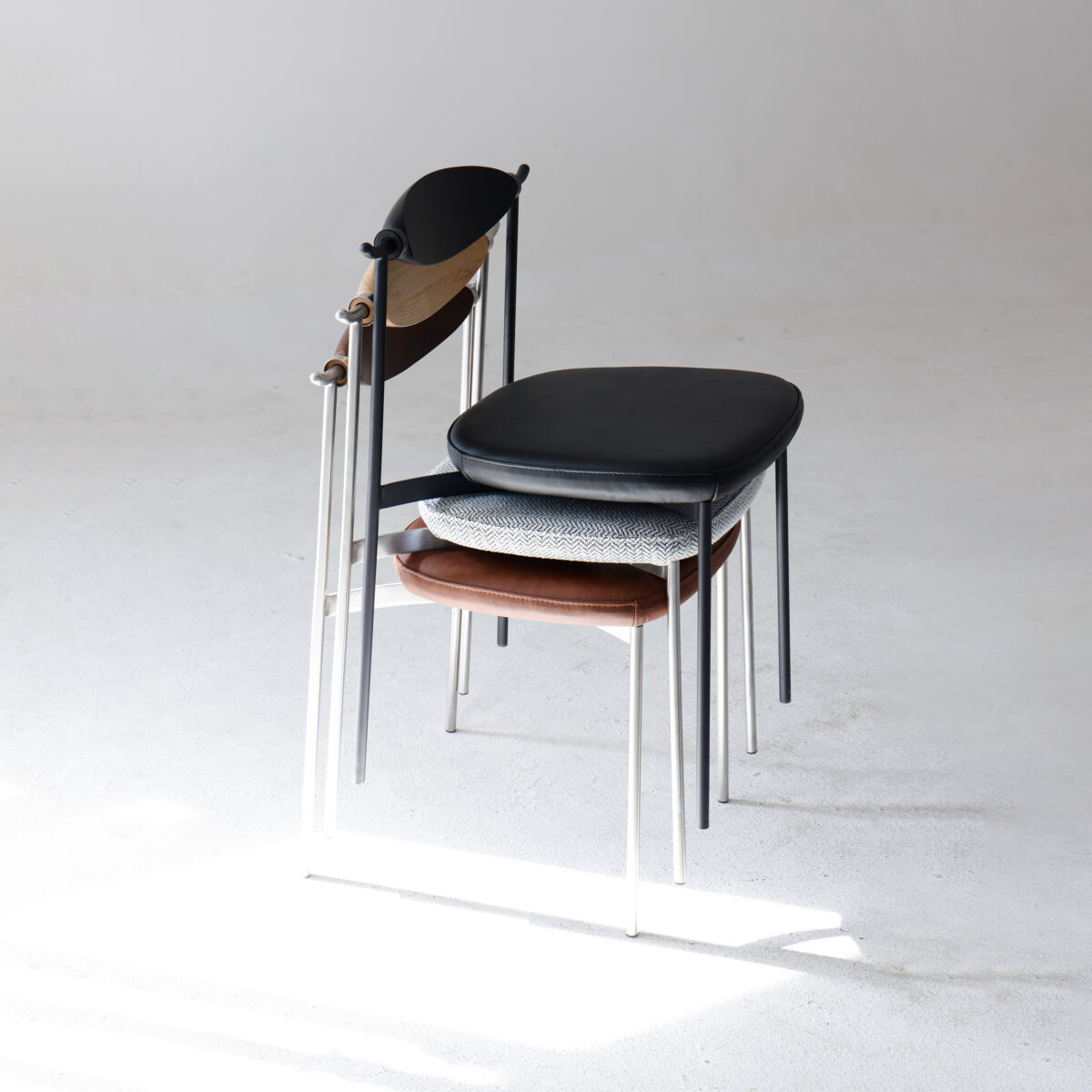 Alison Dining Chair2.0_Black_C2 Three modern chairs with slim metal legs are stacked together on a plain white floor, each chair featuring a differently coloured seat: black, grey patterned, and brown. Soft natural light is cast across the scene.