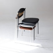 Three modern chairs with slim metal legs are stacked together on a plain white floor, each chair featuring a differently coloured seat: black, grey patterned, and brown. Soft natural light is cast across the scene.