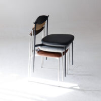 Three modern chairs with slim metal legs are stacked together on a plain white floor, each chair featuring a differently coloured seat: black, grey patterned, and brown. Soft natural light is cast across the scene.