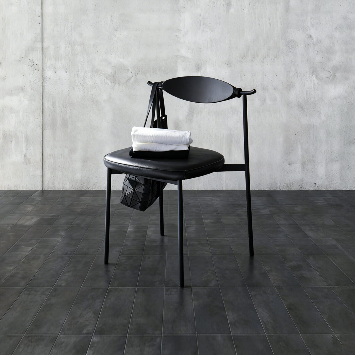 Alison Dining Chair2.0_Black_C5 A modern black chair with a rounded backrest sits on a dark tiled floor. A white folded towel and a black geometric handbag are placed on the seat against a textured grey concrete wall.