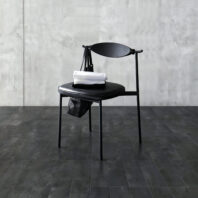 A modern black chair with a rounded backrest sits on a dark tiled floor. A white folded towel and a black geometric handbag are placed on the seat against a textured grey concrete wall.