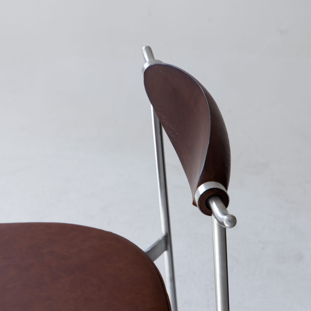 A close-up of a modern chair with a round brown leather seat and a curved wooden backrest, supported by sleek silver metal rods, set against a plain light background.