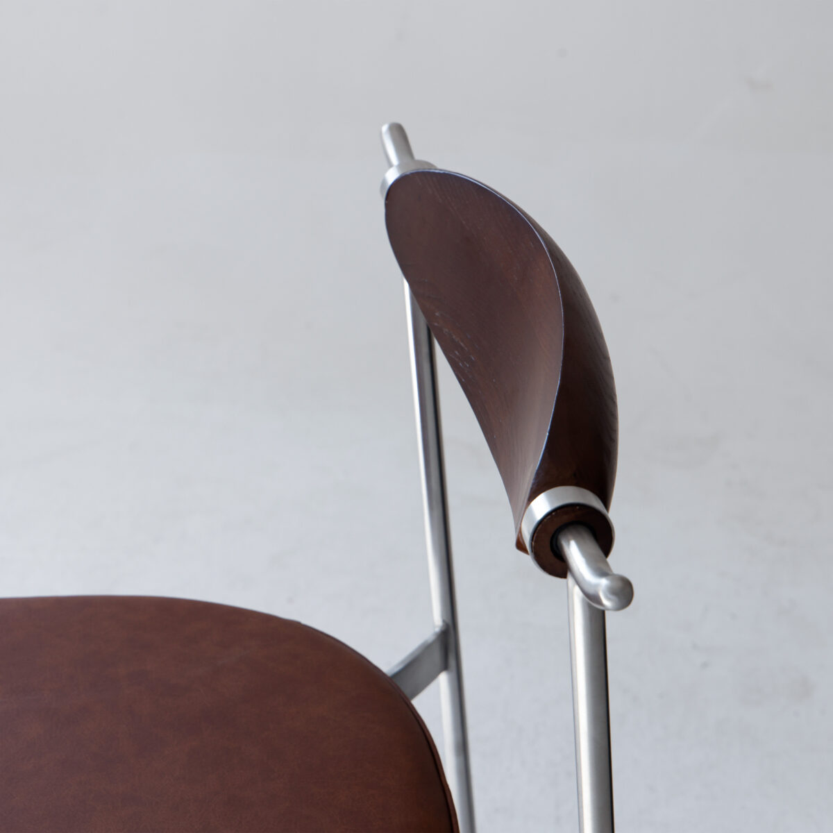 Alison Dining Chair2.0_Dark Brown_Leather_B6 A close-up of a modern chair with a round brown leather seat and a curved wooden backrest, supported by sleek silver metal rods, set against a plain light background.