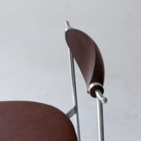 A close-up of a modern chair with a round brown leather seat and a curved wooden backrest, supported by sleek silver metal rods, set against a plain light background.