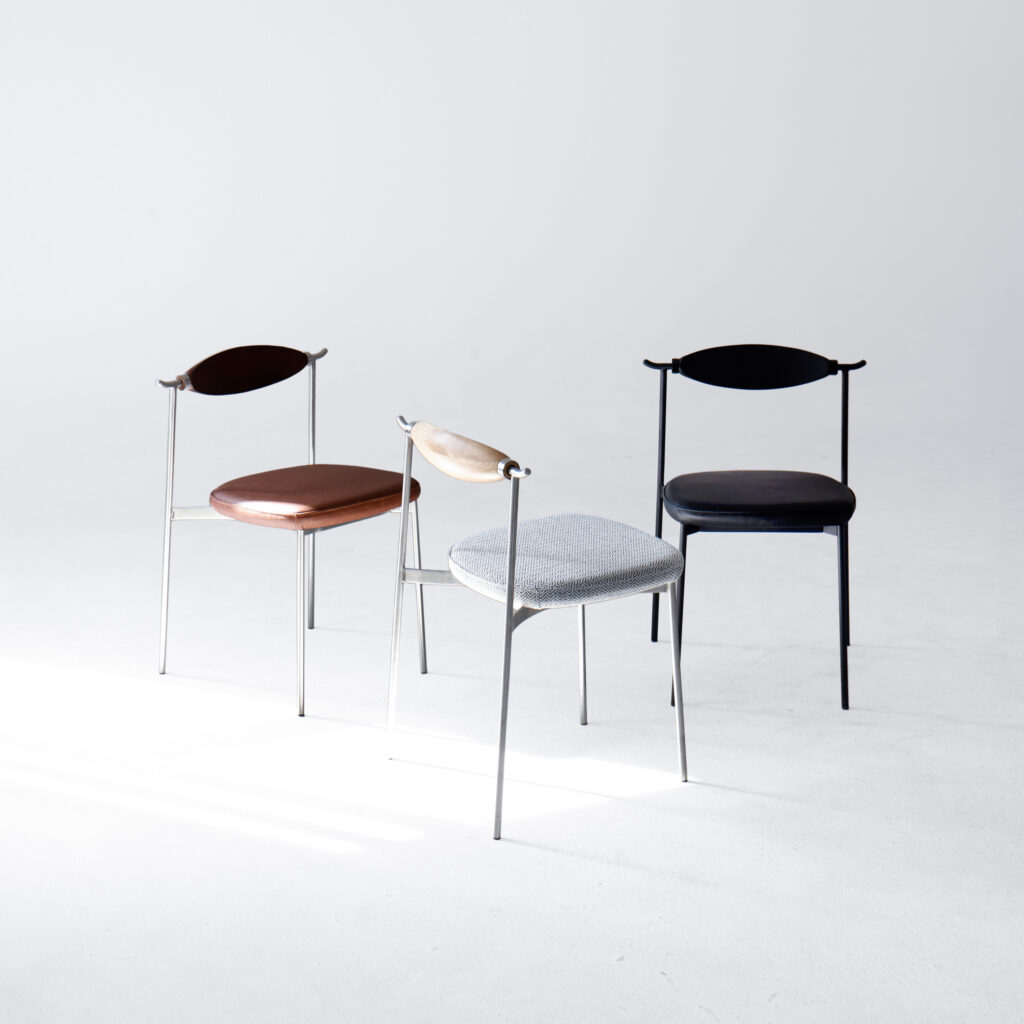 Three minimalist chairs are arranged on a white floor with a plain white background. Each chair has a different seat colourโbrown, light grey, and blackโand slender metal legs, creating a modern and simple aesthetic.