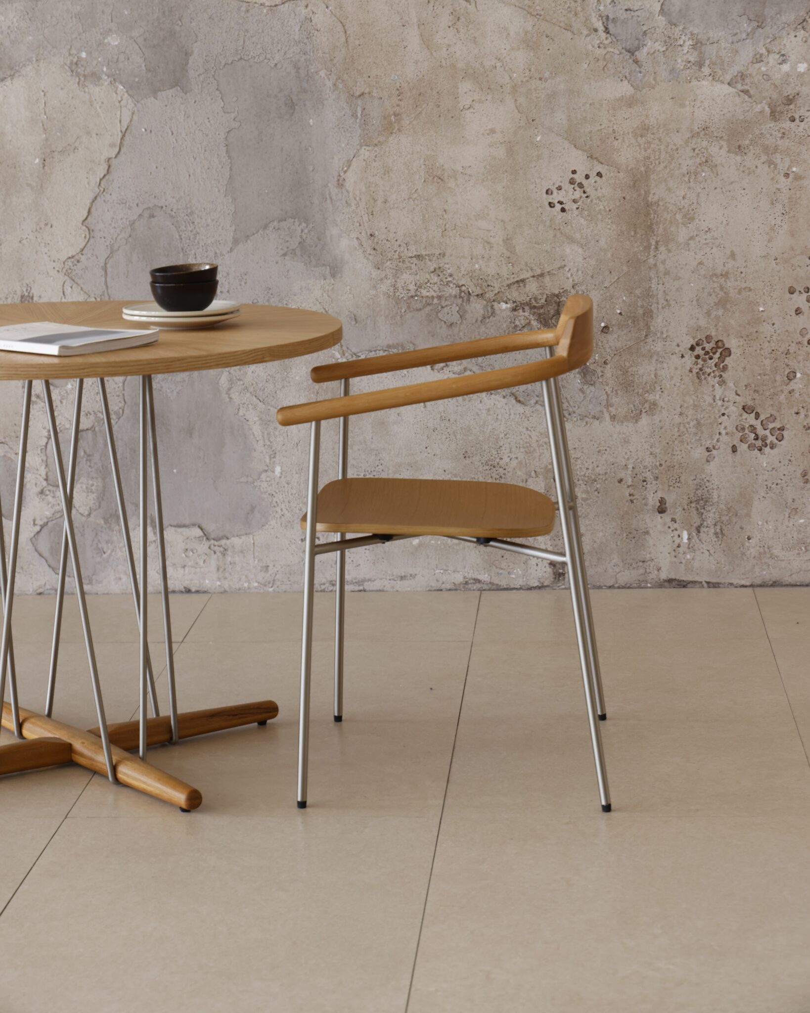 A minimalist scene with a round wooden table, a black cup and saucer, a closed notebook, and a wooden chair with metal legs, set against a textured, weathered wall on a tiled floor.