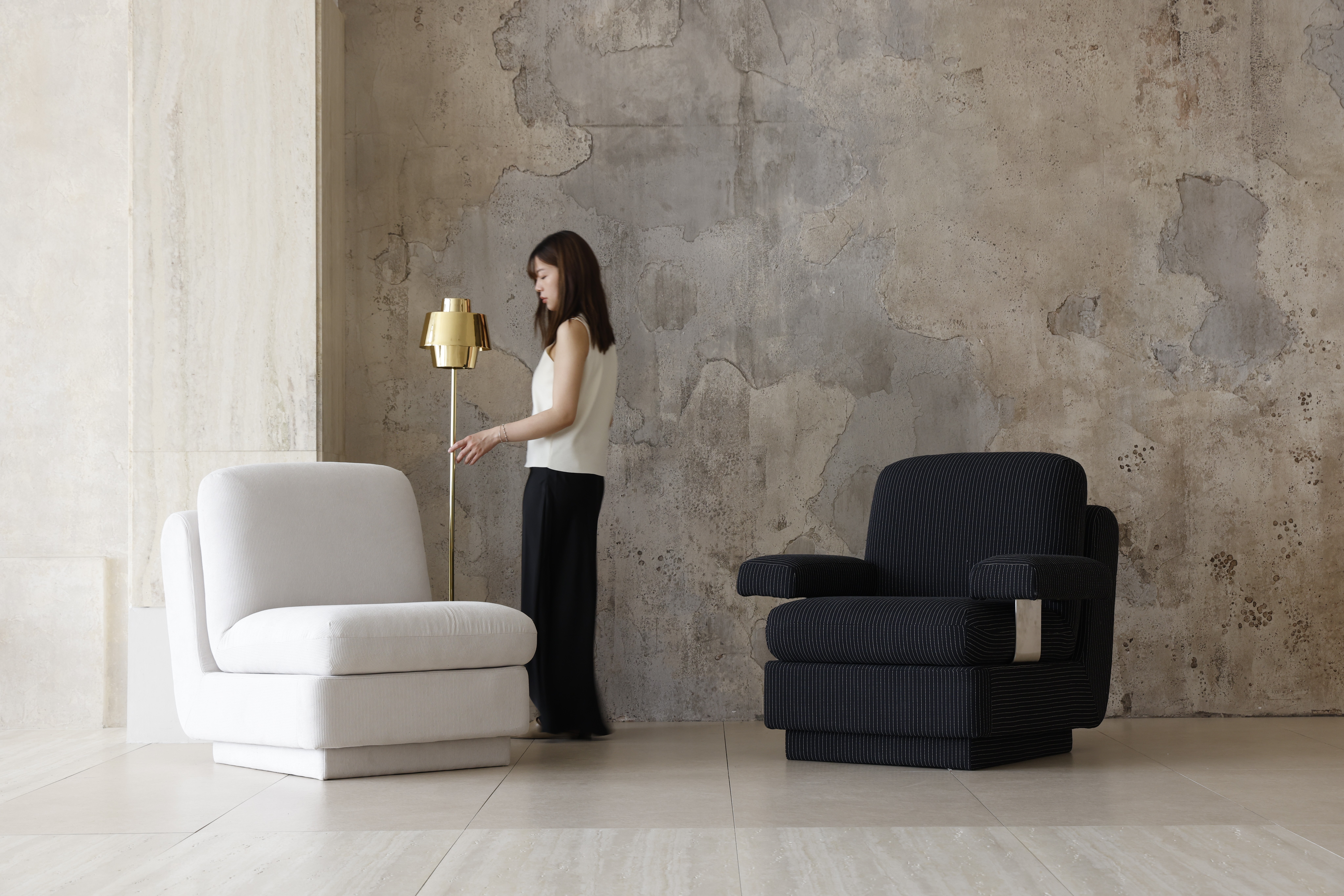 A woman stands near a gold standard lamp between a white chair and a black armchair, in a room with textured, worn concrete walls and light tiled flooring.