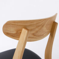 Close-up of the backrest and rear legs of a wooden chair with a natural finish and visible wood grain, featuring a black seat cushion. The background is plain white.