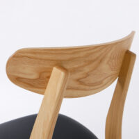 Close-up of the backrest and rear legs of a wooden chair with a natural finish and visible wood grain, featuring a black seat cushion. The background is plain white.