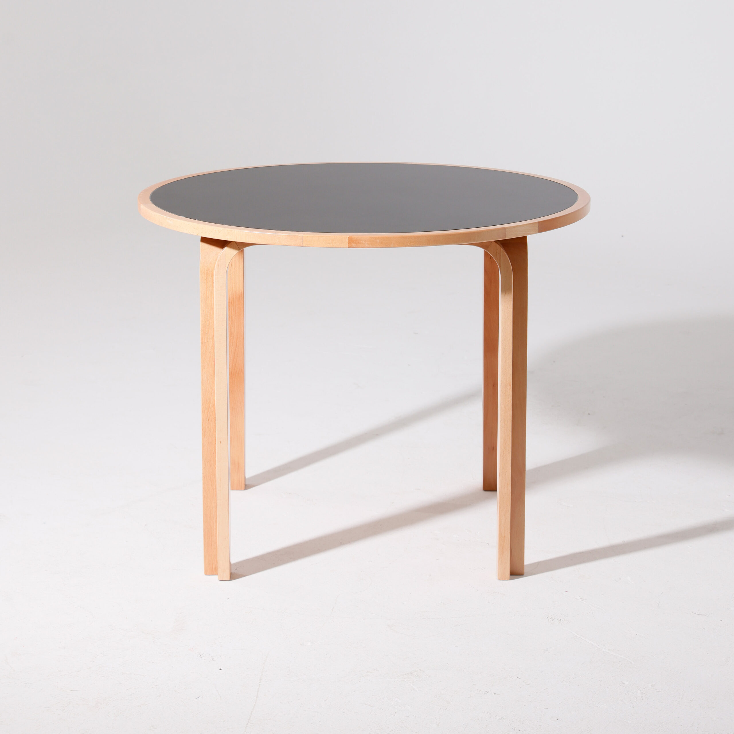 A round table with a light wooden frame and legs, featuring a dark tabletop. The table is set against a plain, light-coloured background.