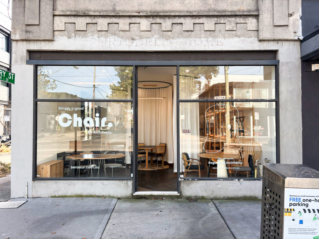 Street view of a modern café named Chai with large glass windows, wooden chairs and tables inside, and a sign on the window reading FREE one-hr parking. Trees and street signs are reflected in the glass.