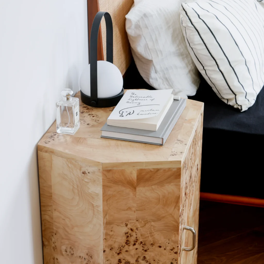 A wooden bedside table with a book, a glass perfume bottle, and a small modern lamp beside a bed with white and striped pillows.